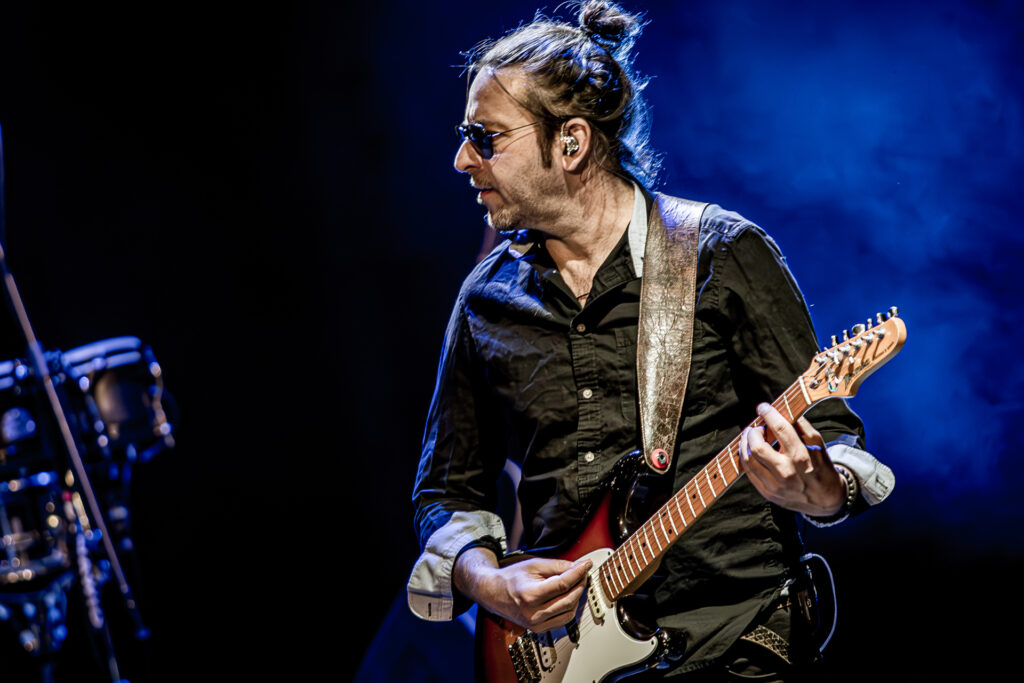 Zdjęcie. Koncert zespołu Sztywny Pal Azji. Mężczyzna w średnim wieku, w ciemnych okularach z dłuższymi włosami gra na gitarze elektrycznej.