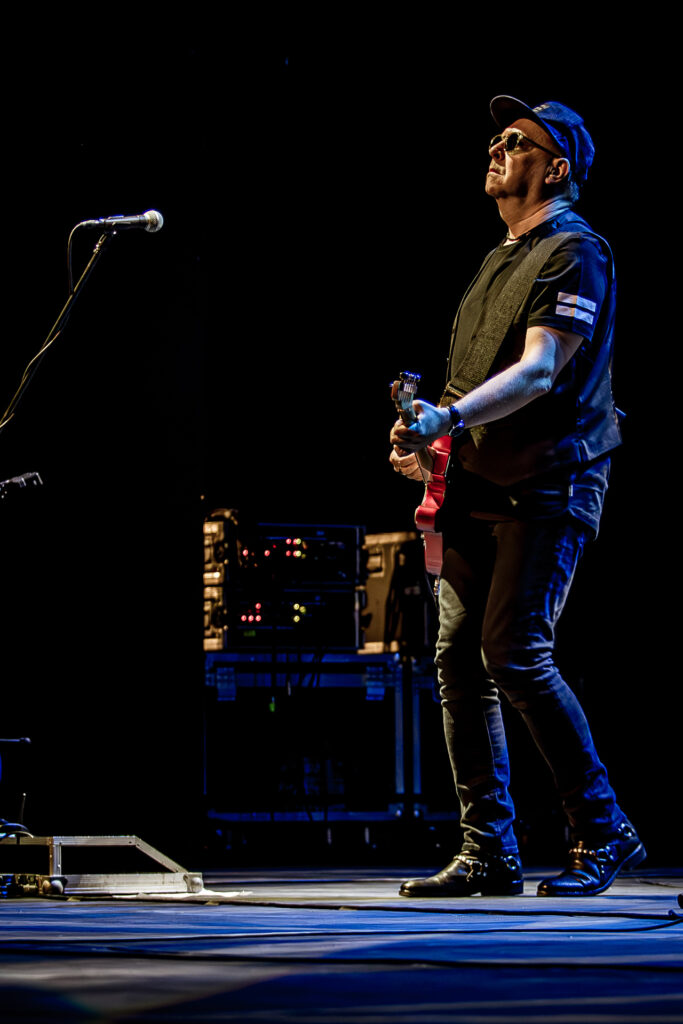 Zdjęcie. Koncert zespołu Sztywny Pal Azji. Mężczyzna w średnim wieku, ciemnych okularach z bejsbolówką na głowie gra na czerwonej gitarze elektrycznej.