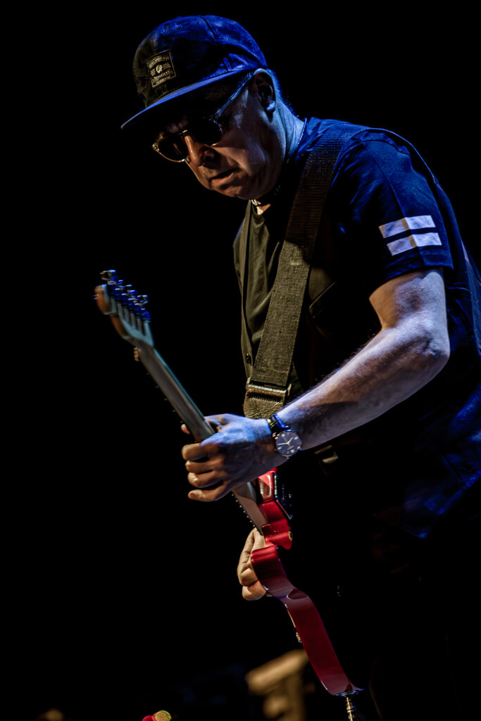 Zdjęcie. Koncert zespołu Sztywny Pal Azji. Mężczyzna w średnim wieku, ciemnych okularach z bejsbolówką na głowie gra na czerwonej gitarze elektrycznej.