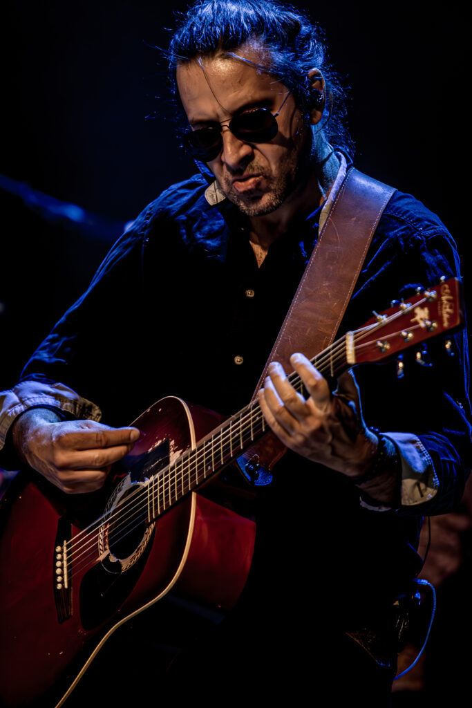 Zdjęcie. Koncert zespołu Sztywny Pal Azji. Mężczyzna w średnim wieku, w ciemnych okularach z dłuższymi włosami gra na gitarze akustycznej.