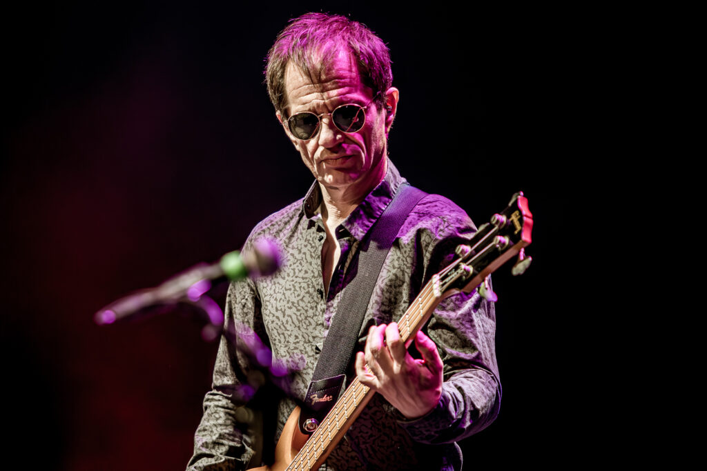 Zdjęcie. Koncert zespołu Sztywny Pal Azji. Szczupły mężczyzna w okularach przeciwsłonecznych gra na gitarze. Oświetla go różowe światło sceniczne.