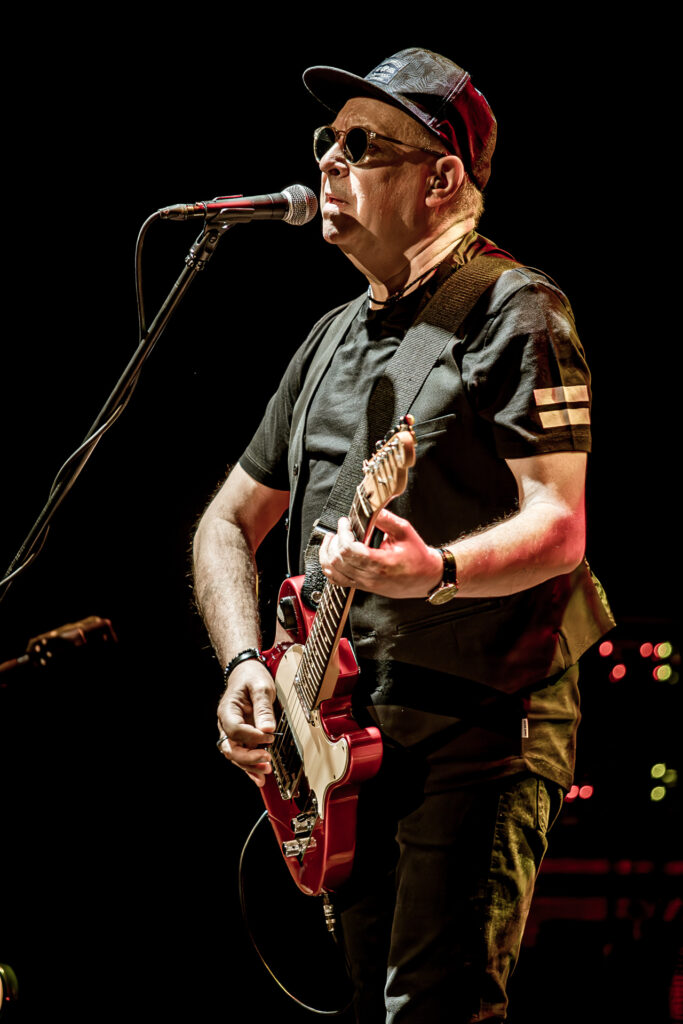 Zdjęcie. Koncert zespołu Sztywny Pal Azji. Mężczyzna w średnim wieku, ciemnych okularach z bejsbolówką na głowie gra na czerwonej gitarze elektrycznej i śpiewa do mikrofonu.