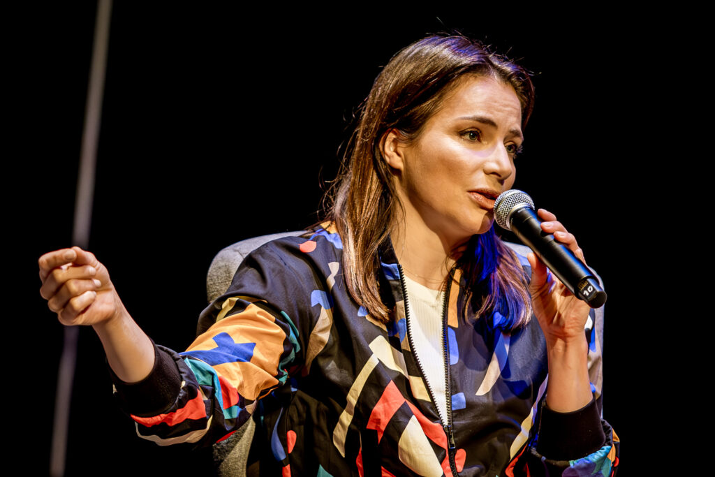 Zdjęcie. Spotkanie z Anną Dereszowską. Kobieta o długich ciemnych wlosach, ubrana we wzorzystą, kolorową kurtkę mówi do mikrofonu. To Anna Dereszowska.