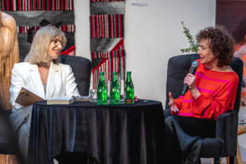 Zdjęcie. Dwie kobiety siedzą przy okrągłym stoliku. To aktorka Joanna Szczepkowska (mówi do mikrofonu) i blondynka w okularach - Aleksandra Niemczyk - prowadząca spotkanie. Obie panie uśmiechają się do siebie.