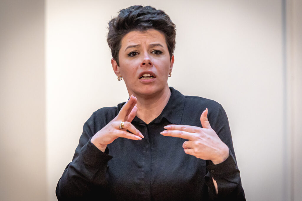 Zdjęcie. Brunetka z kótkimi włosami w czarnej koszuli układa dłonie w gest. To tłumaczka języka migowego Anna Kwiatkowska.