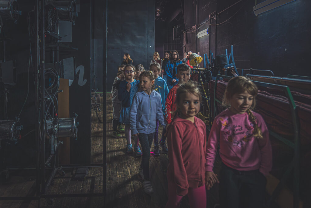 Zdjęcie. Grupa przedszkolaków wędruje w parach kieszenią dużej sceny. Obok nich widoczne meble, reflektory i kable.
