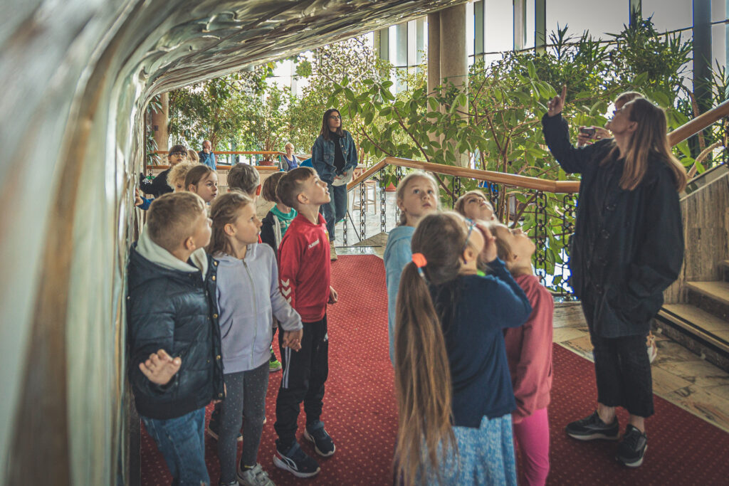 Zdjęcie. Opiekunka wycieczki pokazuje przedszkolakom ozdobną ścianę wyrzeźbioną i pomalowaną przez świętokrzyskiego artystę Adama Wolskiego.