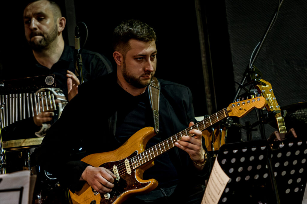 Zdjęcie. Gitarzysta z ciemną brodą w ciemnej marynarce gra na gitarze.