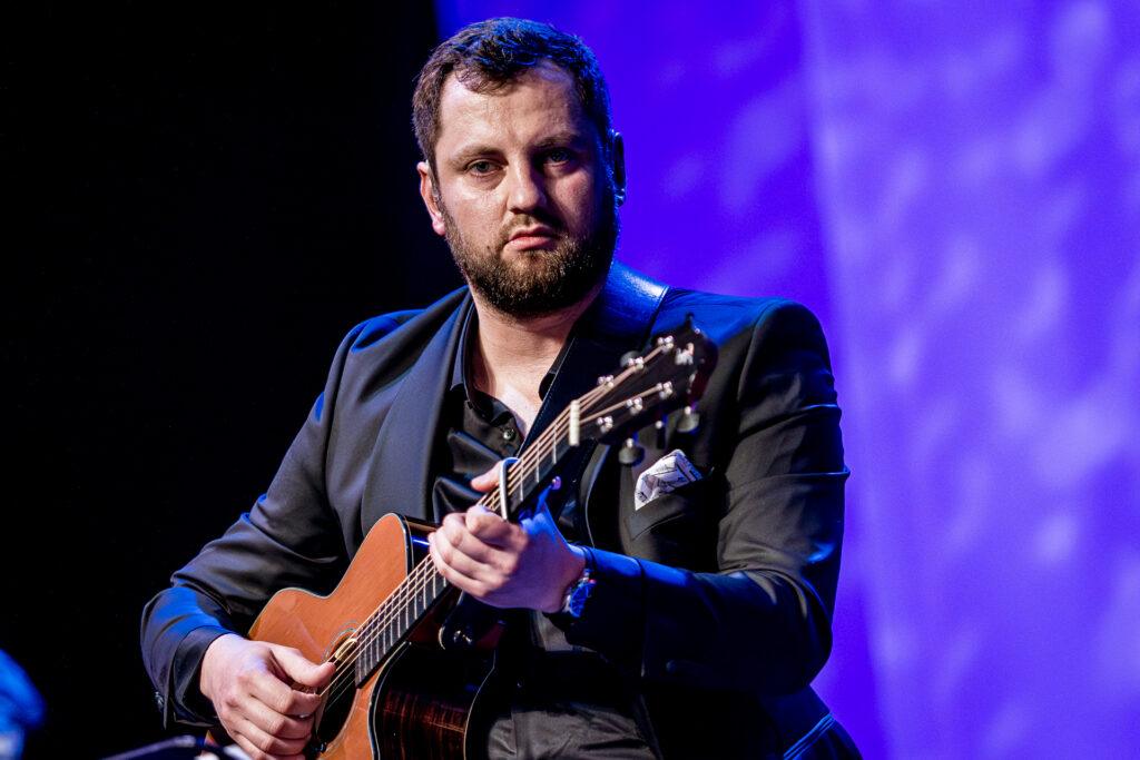 Zdjęcie. Mężczyzna z ciemną brodą w ciemnej marynarce gra na gitarze.