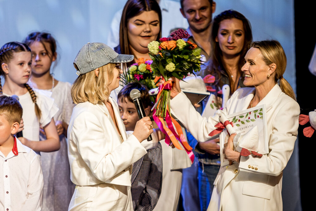 Zdjęcie. Dwie kobiety w białych strojach na scenie KCK. Jedna podajedrugiej kwiaty. W tle uczestnicy koncertu dla Malinki