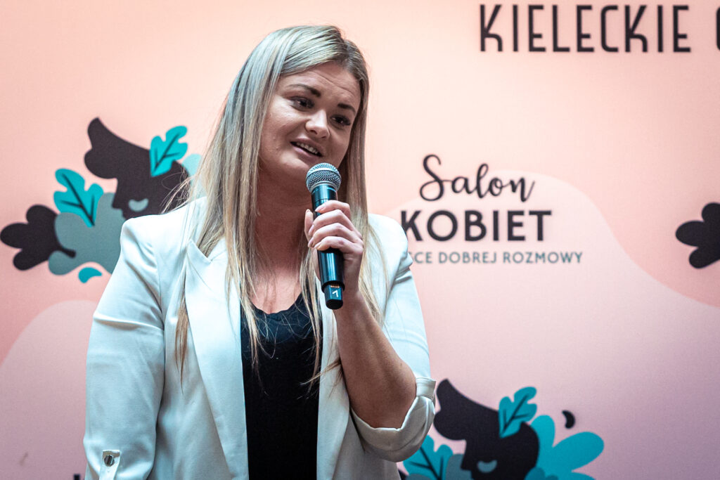 Zdjęcie. Blondynka o długich prostych włosach mówi do mikrofonu. To Aleksandra Bielecka. W tle różowa ścianka z napisem "Salon kobiet".