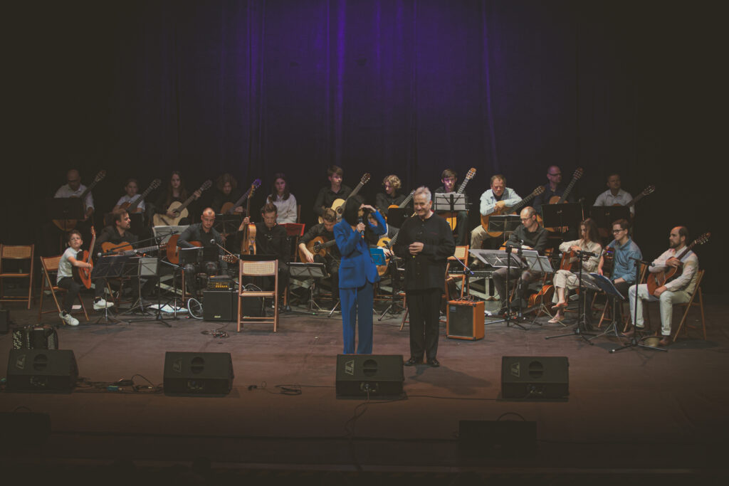 Zdjęcie. Kpobieta w niebieskim kostiumie stoi na scenie. Obok Jerzy Pikor. W tle - gitarowa orkiestra.
