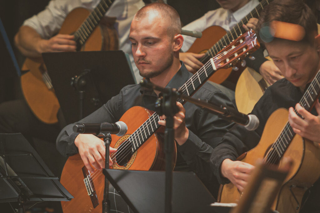 Zdjęcie. Łysawy mężczyzna w ciemnej koszuli gra na gitarze. bok inni gitarzyści.