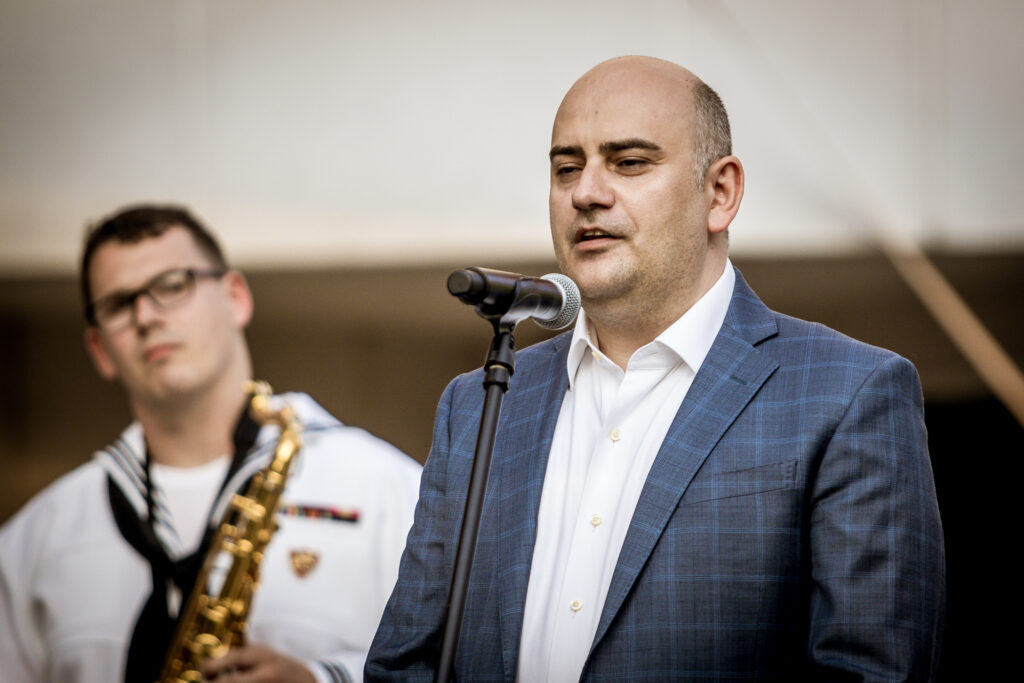 Zdjęcie. Mężczyzna w granatowej marynarce i białej koszuli mówi do mikrofonu. To Erjon Kruja, Konsul ds. Polityczno-Ekonomicznych z Konsulatu USA Krakowie .