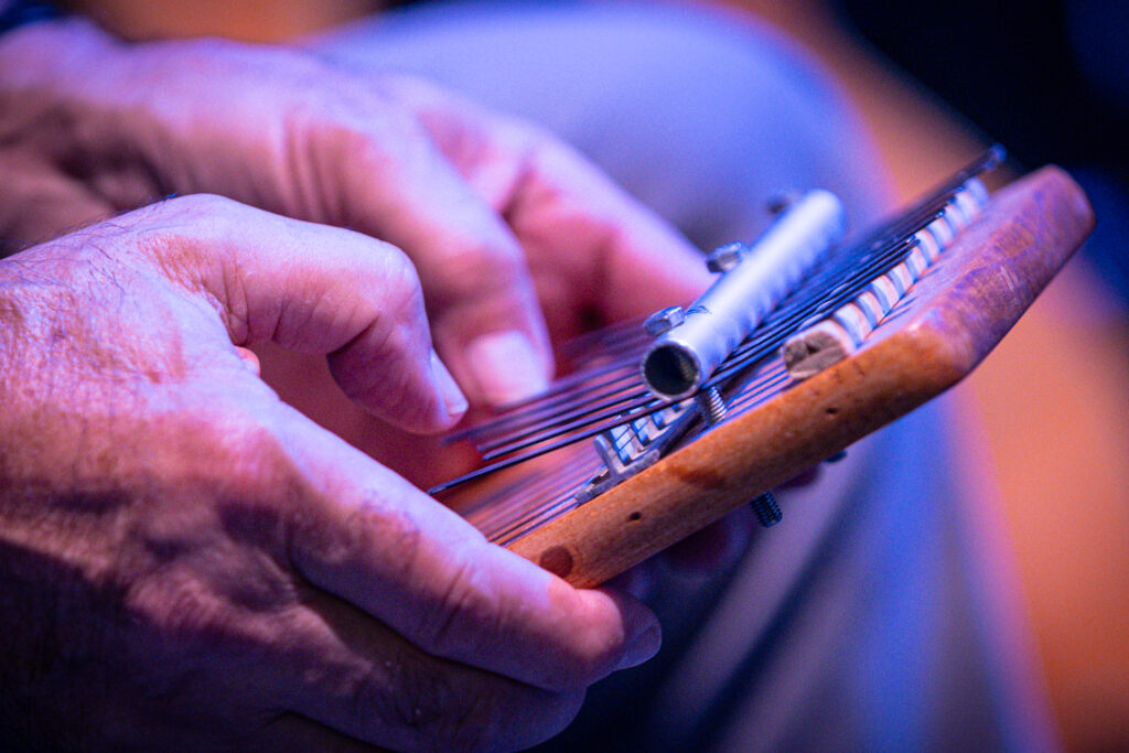 Zdjęcie. Dłonie trzymające niewielki instrument. To kalimba. Na niewielkie j drewnianej płytce ułożone są równolegle cienkie płaskie metalowe blaszki. Dłonie naciskają te pojedyncze blaszki.