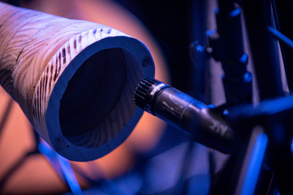 Zdjęcie. Okrągly otwór drewnianej tuby - instrumentu muzycznego przy niewielkim czarnym mikrofonie.