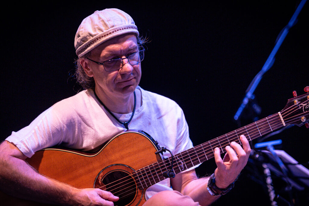 Zdjęcie. Mężczyzna w okularach i czapce gra na gitarze.