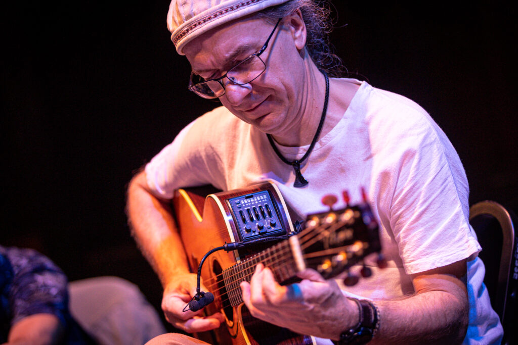 Zdjęcie. Mężczyzna w okularach i czapce gra na gitarze.