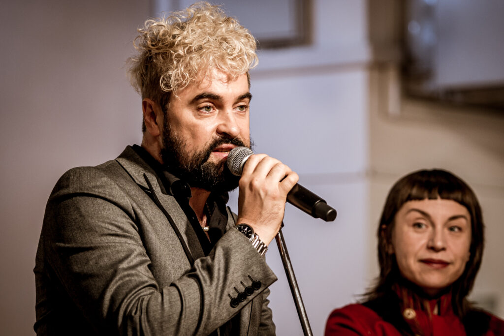 Zdjęci. Mężczyzna o ciemnej brodzie i blond grzywce z lokami mi do mikrofonu. W tle uśmiechnięta brunetka. To Artur Wijata wicedyrektor KCK i artystka Magda Rucińska.