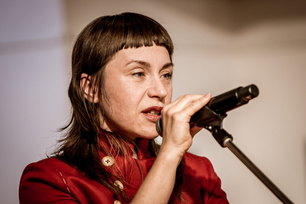 Zdjęcie. Brunetka z prostą krótką grzywką na czole, obrana w czerwoną marynarkę ze złotymi guzikami mówi do mikrofonu. To Magda Rucińska.