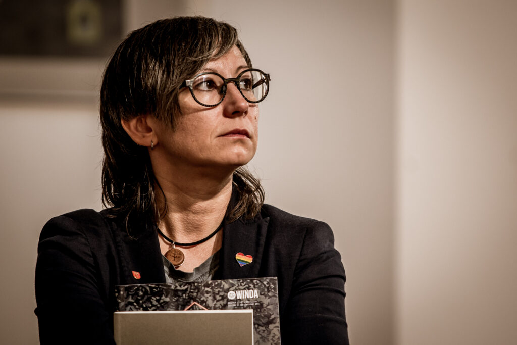 Zdjęcie. Brunetka o włosach do ramion w okularach o ciemnych oprawkach i w czarnej marynarce z tęczowym sercem przyczepionym do klapy patrzy w bok. To radna Katarzyna Zapała.