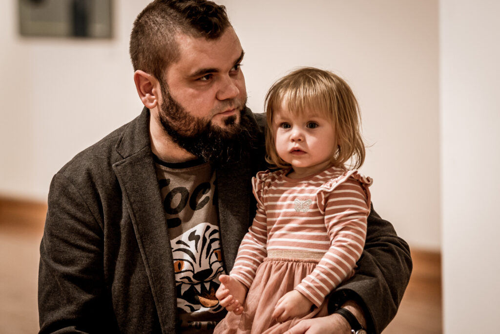 Zdjęcie. Mężczyzna ciemną brodą w brązowej marynarce trzyma na kolanach dwuletnią dziewczynkę o blond włosach ubraną w sukienkę w beżowo białe paski.