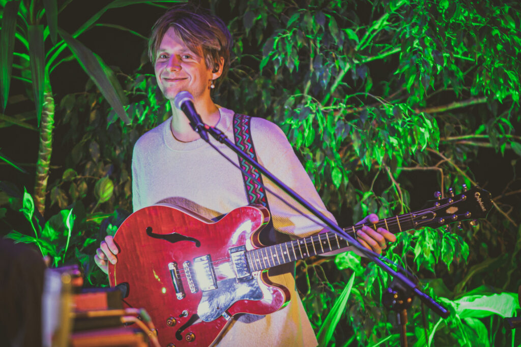 Zdjęcie. Mężczyzna o jasnych włosach z długą grzywką uśmiecha się i gra na gitarze. To muzyk zespołu Muchy. W tle - duże zielone żywe rośliny.