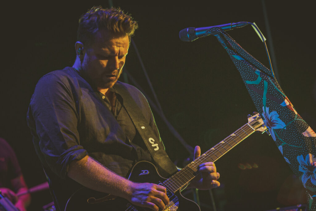 Zdjęcie.Ciemnowłosy mężczyzna z grzywką zaczesaną do góry gra na gitarze. To muzyk zespołu Muchy.