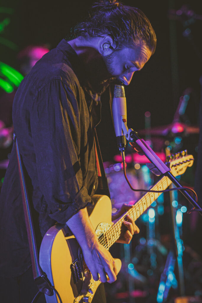Zdjęcie. Mężczyzna o długich włosach spiętych w kucyk gra na żółtej gitarze i śpiewa do mikrofonu.