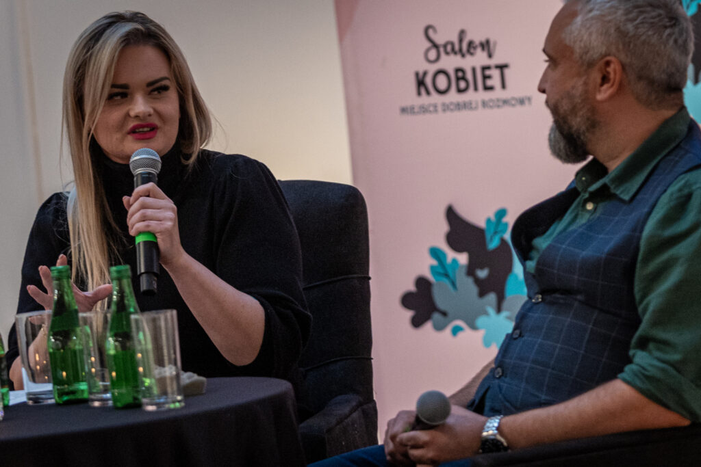 Zdjęcie. Długowłosa kobieta siedzi przy stoliku i mówi coś do mikrofonu. To prowadząca spotkanie Aleksandra Bielecka. Patrzy w kierunku mężczyzny w granatowej kamizelce i zielonej koszuli.