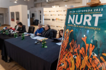 Zdjęcie. Plakat festiwalu NURT stojący na sztaludze. W tle stół przy którym siedzą uczestnicy konferencji prasowej dotyczącej festiwalu.
