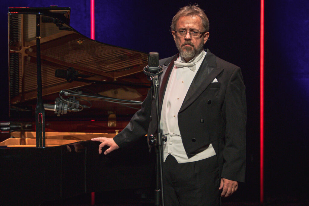 Zdjęcie. Mężczyzna brodą w okularach we fraku stoi przy mikrofonie. Prawą ręką opiera się o fortepian.. To Grzegorz Szostak.