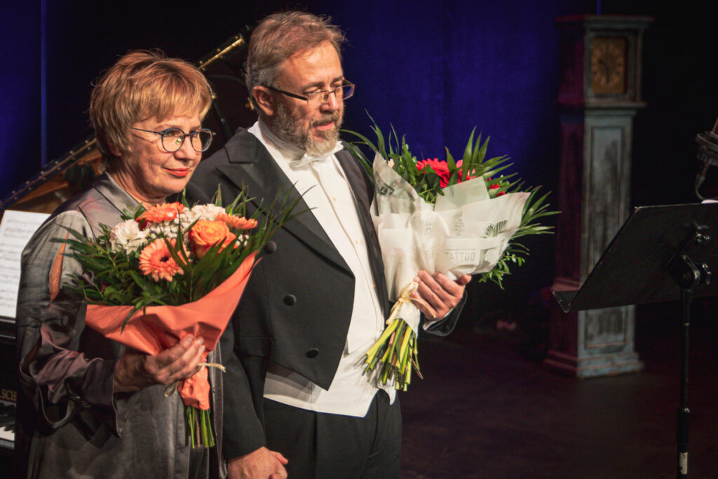 Zdjęcie. Akompaniatorka Danuta Antoszewska i śpiewak operowy Grzegorz Szostak stoją obok siebie na Malej Scenie KCK. W dłoniach trzymają bukiety kwiatów. Uśmiechają się.