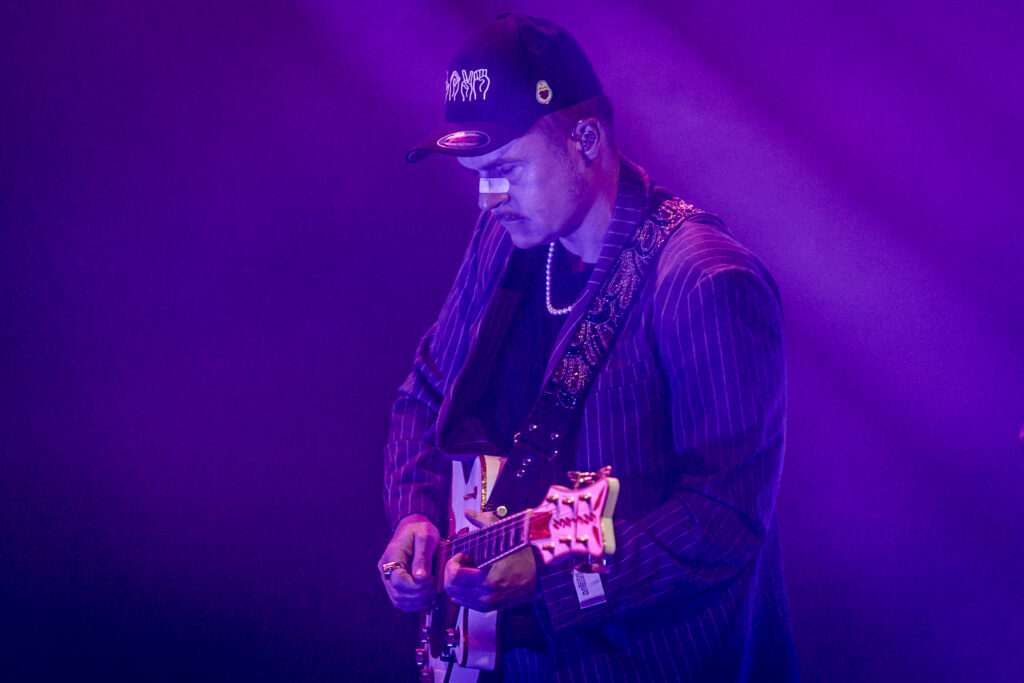 Zdjęcie. Mężczyzna w bejsbolówce i plastrze na nosie gra na gitarze. To Piotr Rogucki oświetlony fioletowym światłem scenicznym.