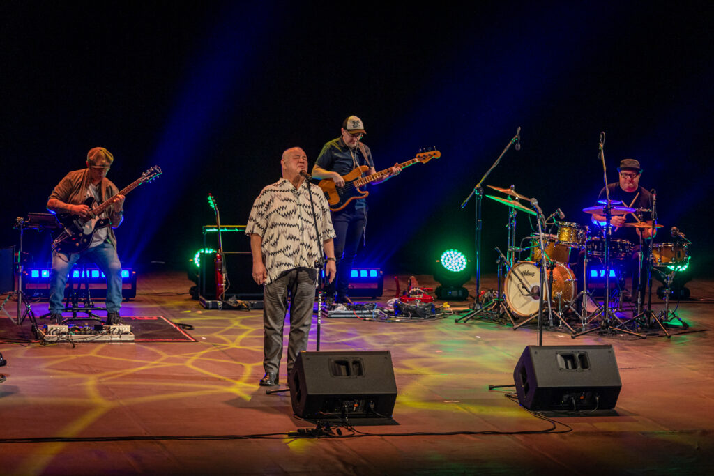Zdjęcie. Stanisław Soyka śpiewa do mikrofonu. W tel dwóch gitarzystów i perkusista.