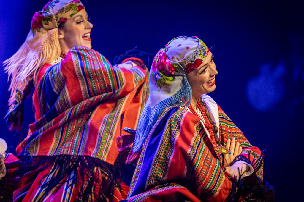 Zdjęcie. Dwie tancerki Mazowsza w jasnych chustach na głowie i pelerynach w kolorowe paski na ramionach.