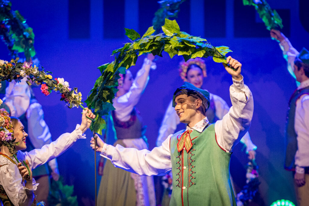 Zdjęcie. Tancerze Mazowsza. Panie i panowie trzymają nad głowami łuki z uplecionych zielonych liści winorośli lub uplecionych kwiatów.