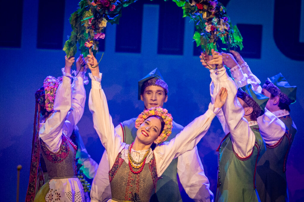 Zdjęcie. Tancerka w żółto-różowym wianku na głowie rozkłada ręce na bok. Nad nią tunel z półokrągłych girland kwiatów, trzymanych przez tancerzy i tancerki stojących w rzędzie. Za tancerką mężczyzna w zielonej czapce rogatywce.