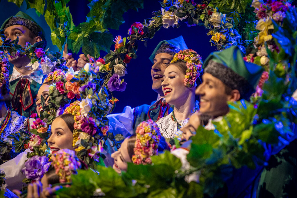 Zdjęcie. Girlandy kwiatów i liści, spomiędzy których wyłaniają się głowy tancerzy: panów w zielonych rogatywkach i pań z użymi opaskami przyozdobionymi kwiatami na głowach. To tancerze zespołu Mazowsze.