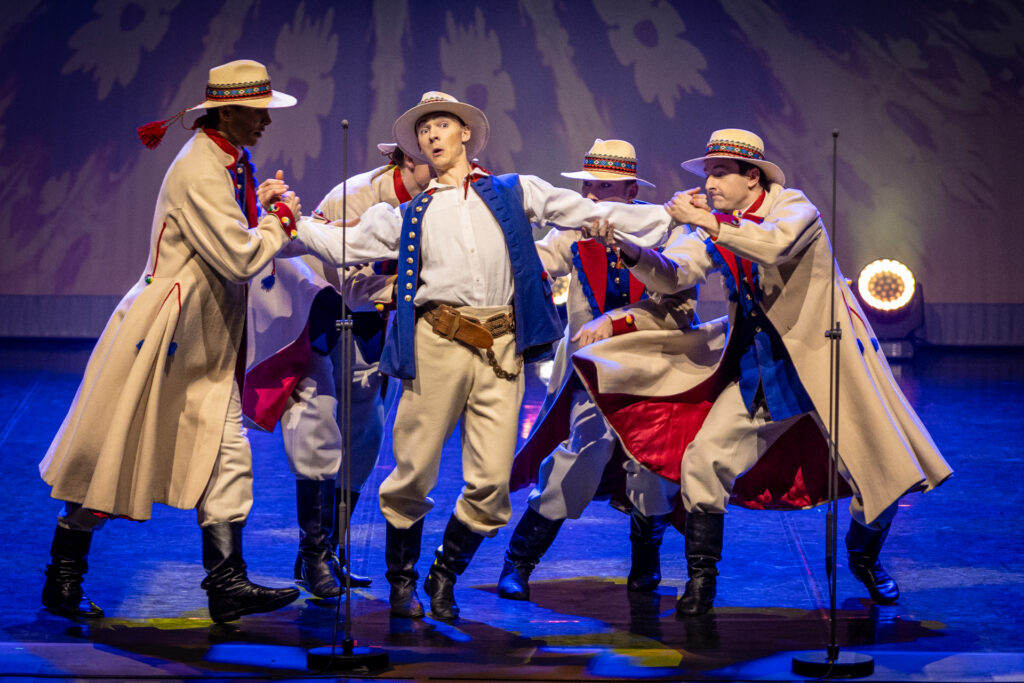 Zdjęcie. Tancerze Mazowsza na scenie. Czterech mężczyzn w kapeluszach i jasnych płaszczak otacza i przytrzymuje piątego, w niebieskiej kamizelce i kapeluszu na głowie.