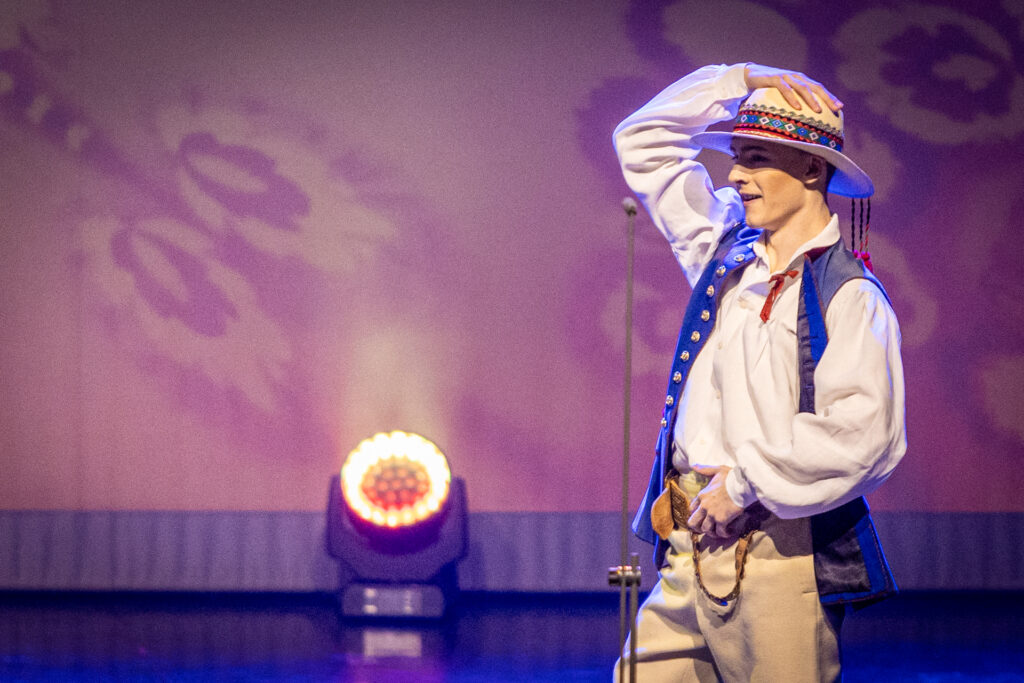 Zdjęcie. Mężczyzna w niebieskiej kamizelce, jasnej koszuli i spodniach stoi na scenie. Prawą ręką trzyma się za kapelusz z rondem i kolorową opaską. Lewą rękę założył za pas. To tancerz Mazowsza w stroju ludowym.