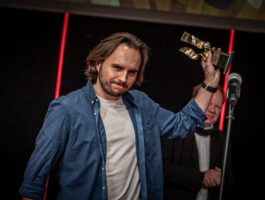 Zdjęcie. Mężczyzna brunet w włosami za ucho, wąsami i brodą trzyma do góry odniesioną statuetkę Nurtu. To zwycięzca tegorocznego festiwalu reżyser Daniel Stopa.