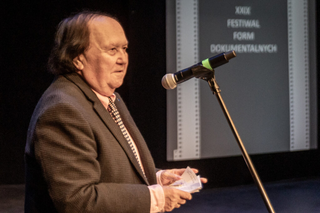 Zdjęcie. Łysawy starszy mężczyzna w ciemnej marynarce mówi do mikrofonu. To dyrektor artystyczny festiwalu Krzysztof Miklaszewski.