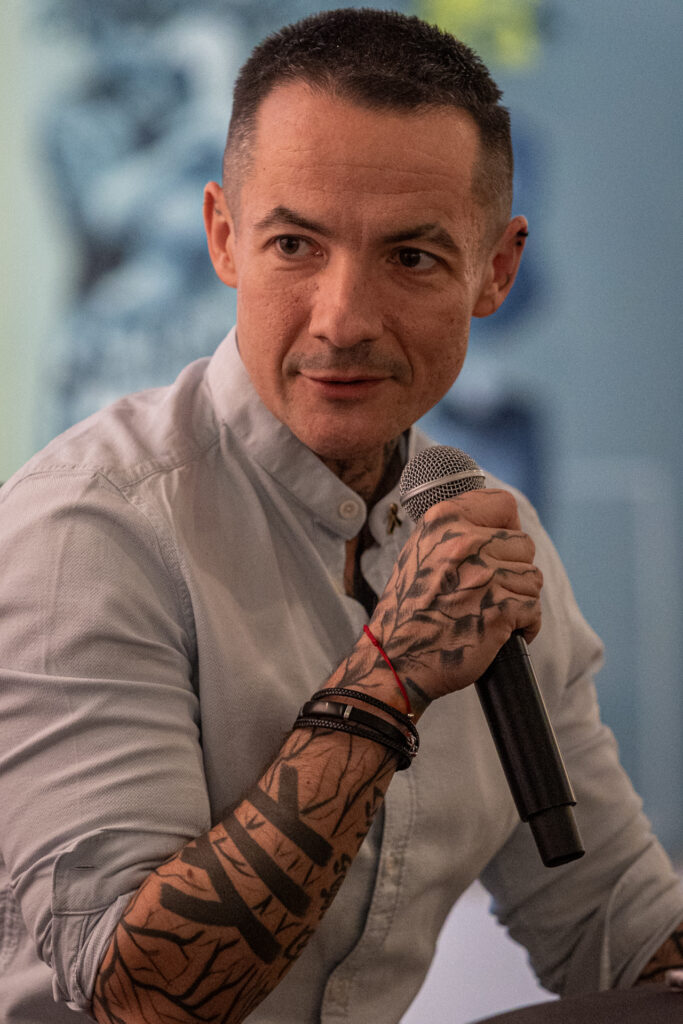 Zdjęcie. Szczupły mężczyzna z wytatuowaną ręką i przedramieniem mówi do mikrofonu. Patrzy w bok. To Marcin Józefaciuk.