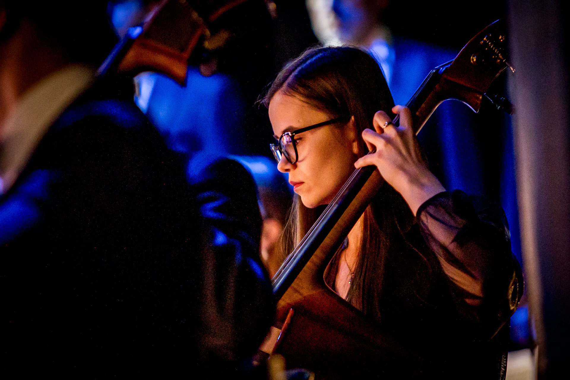 Zdjęcie. Wiolonczelistka o długich prostych włosach i okularach w ciemnych oprawkach gra na instrumencie.