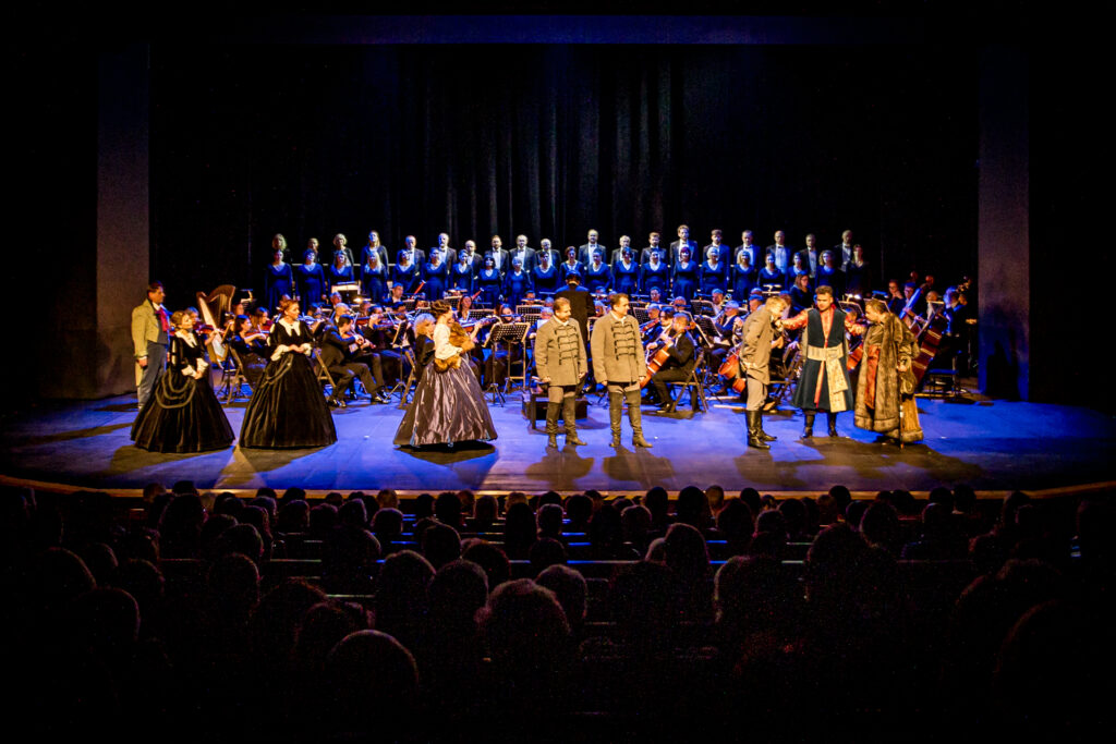 Zdjęcie. Duża Scena KCK. Na niej sześciu mężczyzn i trzy kobiety w sukniach z krynolinami. To soliści opery "Straszny Dwór" w tle orkiestra i chór.