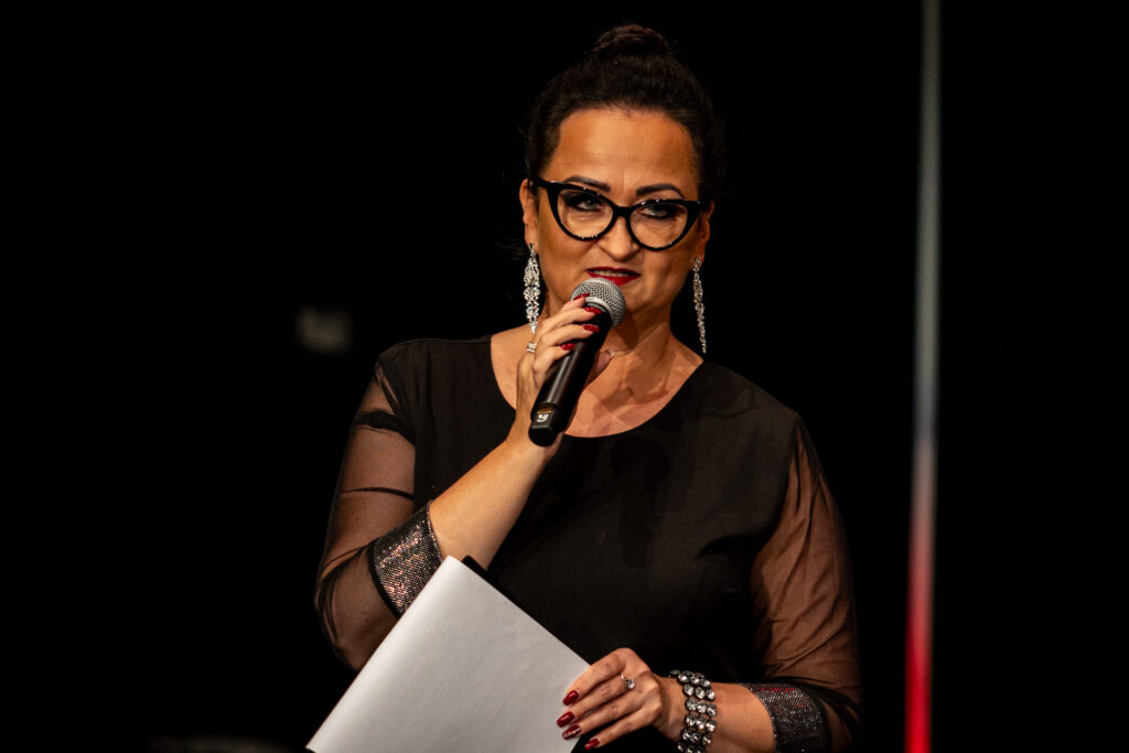 Zdjęcie. Brunetka w czarnej sukni z tiulowymi rękawami mówi do mikrofnu. To zapowiadająca koncert dziennikarka Aneta Czaban.