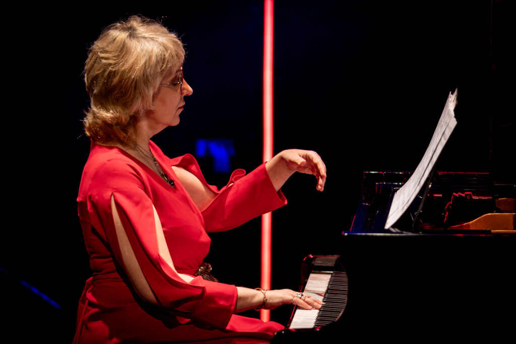 Zdjęcie. Kobieta w czerwonej sukni o włosach blond do ramion gra na fortepianie. To akompaniatorka Ewa Pelwecka.
