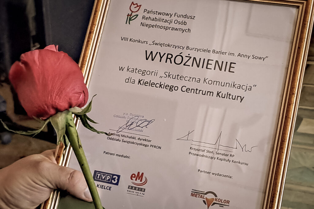 Zdjęcie. Ramka z wyróżnieniem. Na nim napisano: Ośmy konkyrs Burzyciele Barier im. Anny Sowy. Wyróżnienie w kategorii "Skutezna komunikacja" dla Kieleckiego Centrum Kultury.