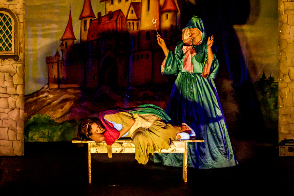 Zdjęcie. Kobieta w zielonej sukni z kapturem załozónym na głowę i różdżką stoi nad Kopciuszkiem śpiącym na ławce.